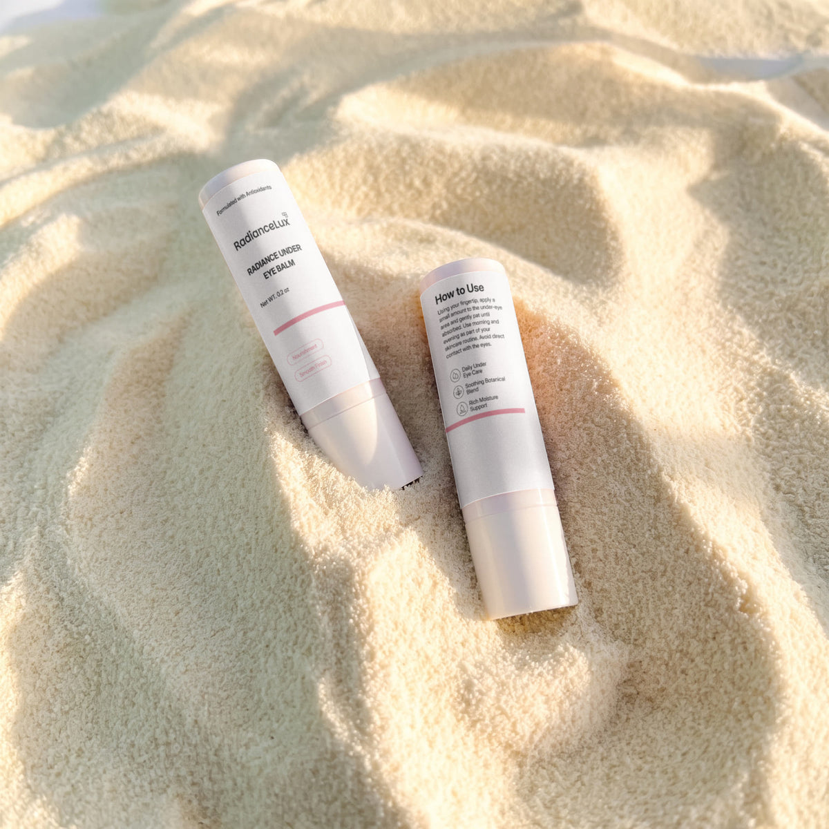 Two Radiance Under Eye Balm sticks, one uncapped, resting on soft sand