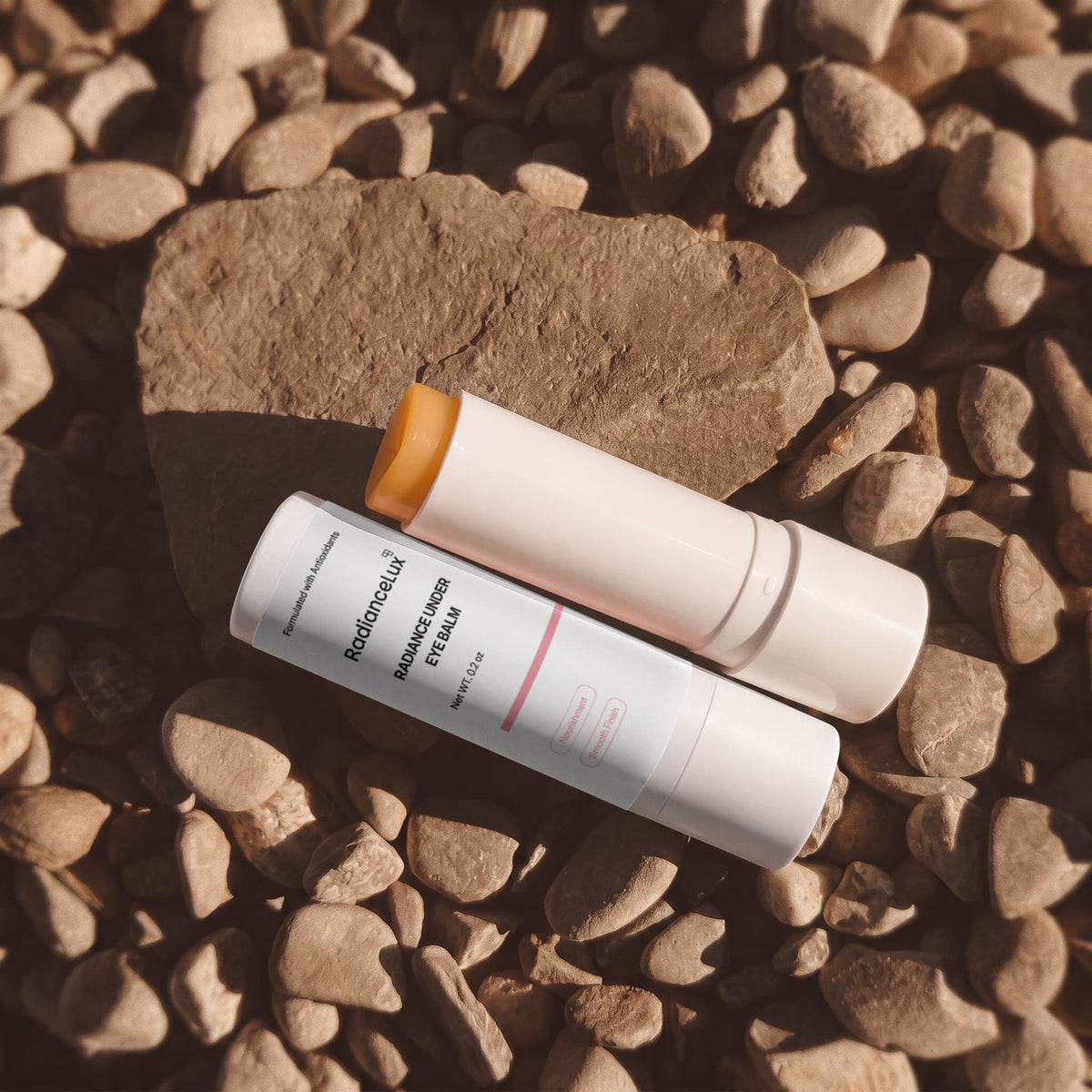 Two under eye balm sticks, one open, lying on brown river stones