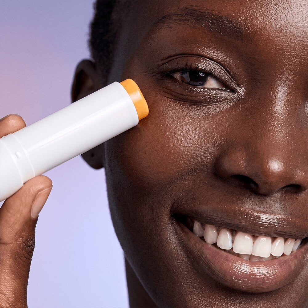 Smiling woman holding the open Radiance Under Eye Balm stick beside her eye