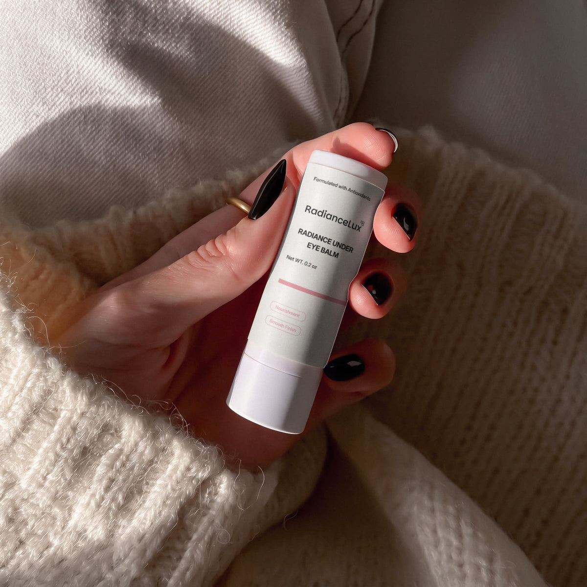 Hands with dark nail polish holding the Radiance Under Eye Balm stick over a cream knit sweater