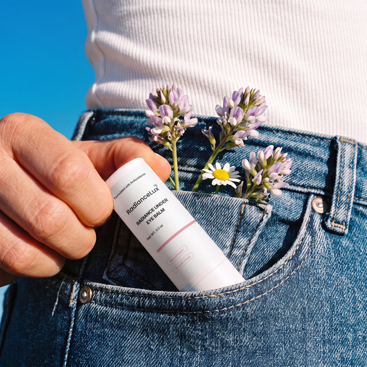 Radiance Under Eye Balm stick tucked in a jeans pocket with small purple flowers and a daisy